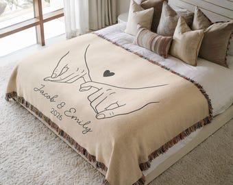 May include: A beige woven throw blanket featuring a black line art illustration of two hands pinky promising with a small heart. The text "Jacob & Emily" and "2026" is printed in black script. The blanket has a multicolored fringed edge.