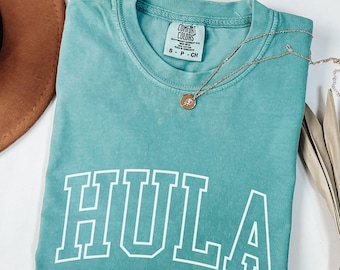 May include: A seafoam green short-sleeve t-shirt with a crew neckline and the word HULA printed in large white block letters across the chest. The shirt features a Comfort Colors brand label at the inner neck and a thin gold-toned chain necklace.