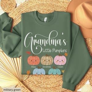 May include: A military green long-sleeve sweatshirt featuring the white text Grandma's Little Pumpkins above five illustrated pumpkins in orange, tan, pink, light blue, and teal, each with a smiling face and a name printed underneath.