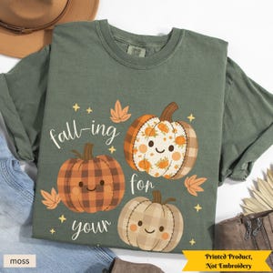 May include: Moss green t-shirt with a fall-themed graphic. The design features three smiling pumpkins with plaid and floral patterns, surrounded by leaves and stars. The text "fall-ing for your" is above the pumpkins. The bottom right corner reads "Printed Product, Not Embroidery".