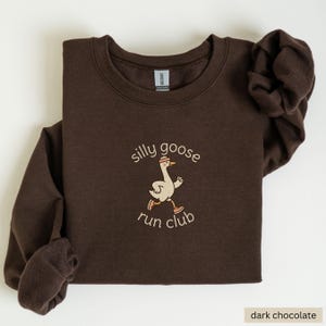 May include: Dark chocolate-colored sweatshirt with the text "silly goose run club" and an embroidered graphic of a running goose wearing a headband and sneakers. The sweatshirt is made of a soft material.