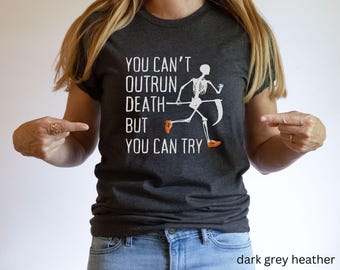May include: Dark grey heather t-shirt with the text "YOU CAN'T OUTRUN DEATH - BUT YOU CAN TRY" and a white skeleton graphic running with a scythe. The skeleton is wearing orange shoes. The shirt is a crew neck.