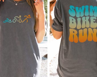 May include: A dark gray t-shirt with a graphic of a swimmer, a cyclist, and a runner. The back of the shirt has the words "Swim Bike Run" in a retro font with a gradient of blue, yellow, and orange.