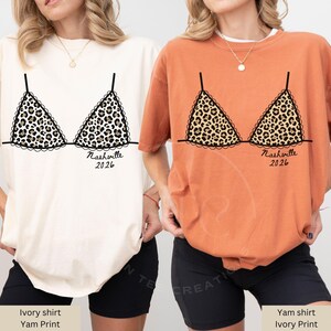 May include: Two t-shirts, one ivory and one yam-colored, each featuring a leopard print bra design. The design includes the text "Nashville 2026" in black script. The shirts are casual and comfortable.