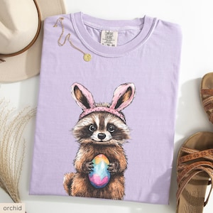 May include: A purple t-shirt with a cartoon raccoon wearing bunny ears and holding a colorful Easter egg. The text "COMFORT COLORS" is on the tag.