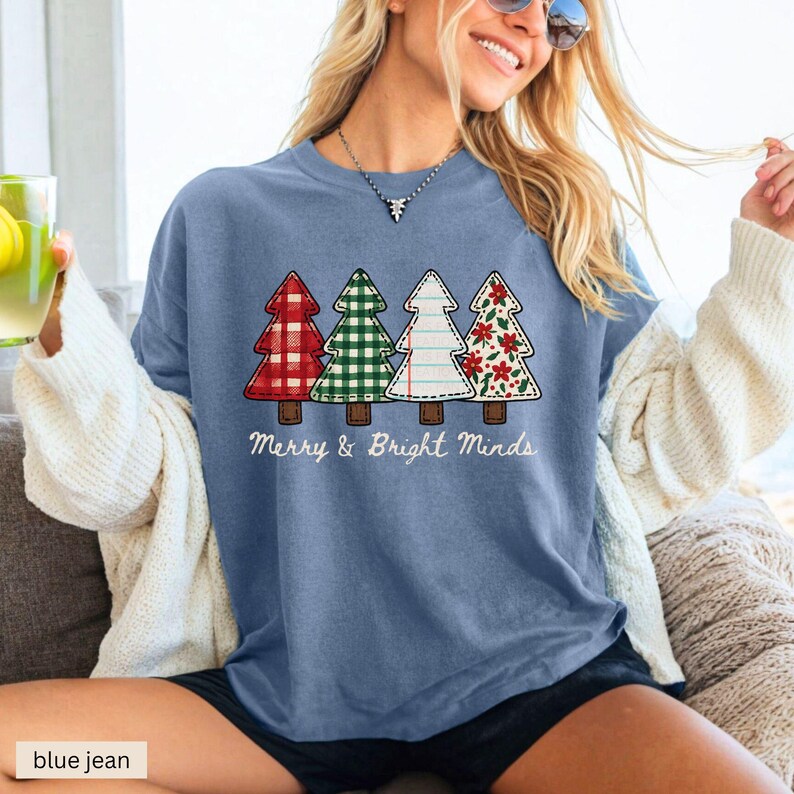 May include: A blue jean colored t-shirt featuring four Christmas trees in red plaid, green gingham, white with stitching, and red floral patterns. The text "Merry & Bright Minds" is printed below the trees. The shirt is a casual, festive design.