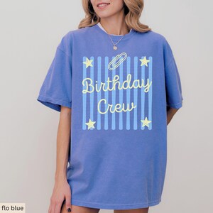 May include: A periwinkle blue short-sleeve t-shirt featuring a central graphic of vertical light blue stripes, four yellow stars, a yellow paperclip icon, and the yellow text "Birthday Crew" written in a cursive script font.
