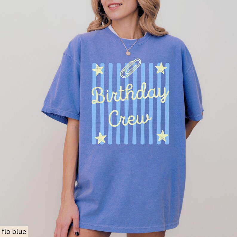 May include: A periwinkle blue short-sleeve t-shirt featuring a central graphic of vertical light blue stripes, four yellow stars, a yellow paperclip icon, and the yellow text "Birthday Crew" written in a cursive script font.