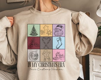 Op de afbeelding: Zandkleurige sweatshirt met een raster van kerstthema-illustraties, waaronder een boom, de kerstman en een peperkoekmannetje. De tekst "IN MY CHRISTMAS ERA" staat eronder gedrukt, met "Your Custom's Version".