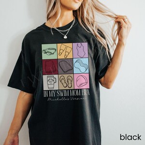 May include: A black t-shirt featuring a grid of nine colorful squares containing line art icons of swimming gear, including goggles, fins, a swimsuit, a towel, a kickboard, a noodle, a tumbler, flip-flops, and a cap, with text reading IN MY SWIM MOM ERA and Michelle's Version.