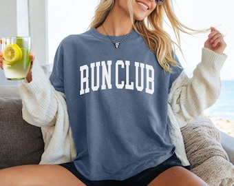 May include: A woman wearing a blue jean shirt with the text "RUN CLUB" in white letters. She is wearing black shorts and a white cardigan.