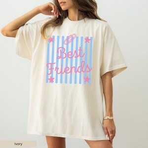 May include: An ivory short-sleeved t-shirt featuring a central graphic of vertical light blue stripes. The design includes the pink text "Best Friends" in a cursive font, a pink paperclip icon at the top, and four pink stars at the corners.