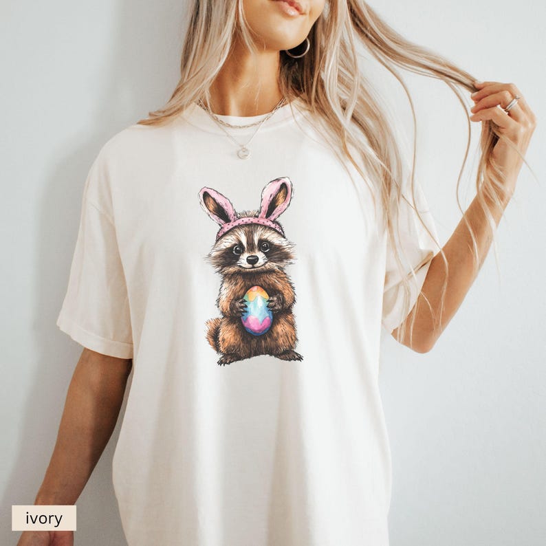 May include: A white t-shirt with a graphic of a raccoon wearing pink bunny ears and holding a colorful Easter egg. The text "ivory" is visible in the bottom left corner of the image.