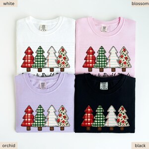 May include: Four folded t-shirts in white, pink, orchid, and black. Each shirt features a Christmas tree design with red plaid, green gingham, white lined, and floral patterns. The text "Merry & Bright" is below the trees.