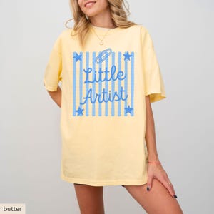 May include: A butter yellow short-sleeve t-shirt featuring a light blue graphic print. The design includes vertical stripes, four stars, a paperclip icon, and the text "Little Artist" in a cursive script centered on the front of the garment.