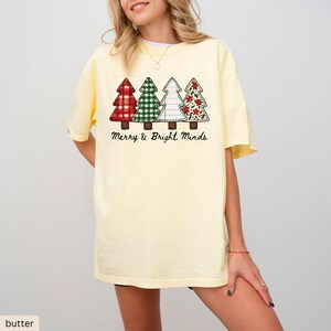 May include: Butter yellow t-shirt featuring four Christmas tree designs in red, green, white, and red with floral patterns. The text "Merry & Bright Minds" is printed below the trees. The shirt has short sleeves and a relaxed fit.
