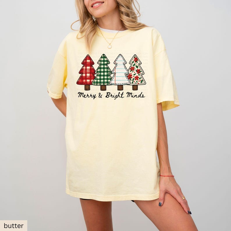 May include: Butter yellow t-shirt featuring four Christmas tree designs in red, green, white, and red with floral patterns. The text "Merry & Bright Minds" is printed below the trees. The shirt has short sleeves and a relaxed fit.