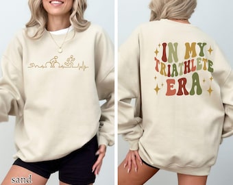 May include: A beige sweatshirt with a graphic of a person riding a bicycle, running, and a heartbeat on the front. The back of the sweatshirt has the text "In My Triathlete Era" in a retro font with colourful accents.