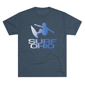 May include: A navy blue t-shirt featuring a surfer in a blue gradient design, riding a surfboard. The text "SURF OHIO" is printed below the surfer in a white and blue gradient. The shirt is made of a soft material.