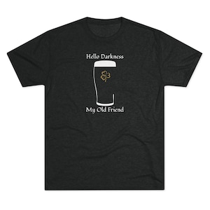 May include: Dark gray t-shirt with the text "Hello Darkness My Old Friend." A white outline of a beer glass is in the center, with a gold shamrock inside. The shirt is made of a soft material.