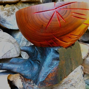 May include: A hand-carved wooden bowl with a smooth, reddish-brown finish and visible grain patterns. The bowl features deep, angular grooves carved into its exterior surface and rests upon a dark, textured, irregular wooden base.