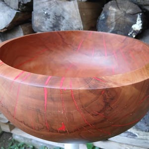 Op de afbeelding: Een ronde houten kom met een gladde, gepolijste afwerking en natuurlijke bruine houtnerven. De kom is geaccentueerd met levendige, dunne, verticale strepen van felrode hars die de natuurlijke scheuren in het houtoppervlak opvullen.