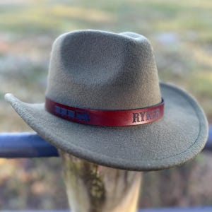 May include: A gray fedora-style hat with a dark red leather band. The band features the word "RYKER" in capital letters. The hat is resting on a wooden post, with a blurred outdoor background.
