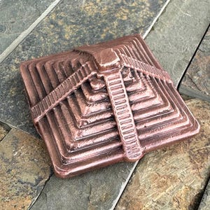 K&ouml;nnte beinhalten: Ein bronzefarbenes, quadratisches Deko-Objekt mit einem Stufenpyramiden-Design. Das Objekt weist ein zentrales vertikales Element auf, das einer Leiter &auml;hnelt, sowie horizontale Linien. Die Pyramide hat mehrere Schichten und besteht aus Metall.