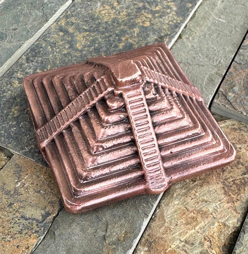 K&ouml;nnte beinhalten: Ein bronzefarbenes, quadratisches Deko-Objekt mit einem Stufenpyramiden-Design. Das Objekt weist ein zentrales vertikales Element auf, das einer Leiter &auml;hnelt, sowie horizontale Linien. Die Pyramide hat mehrere Schichten und besteht aus Metall.