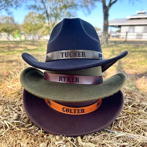May include: Three cowboy hats stacked on top of each other. The top hat is navy blue with a brown band that reads "TUCKER". The middle hat is olive green with a brown band that reads "RYKER". The bottom hat is dark brown with an orange band that reads "COLTER".
