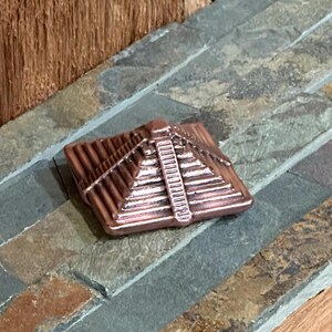 K&ouml;nnte beinhalten: Ein kupferfarbenes, pyramidenf&ouml;rmiges Dekorationsobjekt. Die Pyramide hat detaillierte horizontale Linien und eine vertikale Linie in der Mitte. Das Objekt liegt auf einer mehrfarbigen Steinoberfl&auml;che.