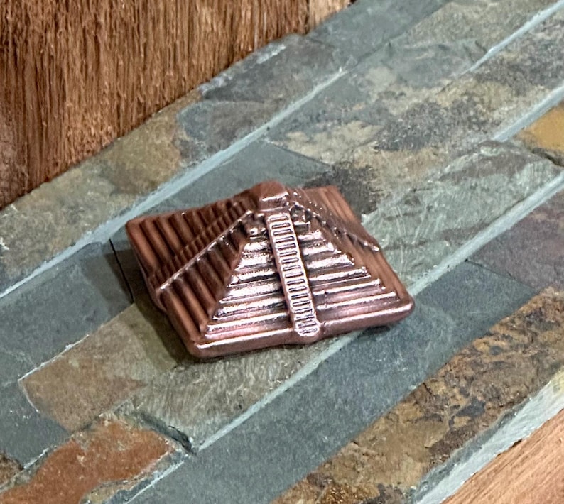 K&ouml;nnte beinhalten: Ein kupferfarbenes, pyramidenf&ouml;rmiges Dekorationsobjekt. Die Pyramide hat detaillierte horizontale Linien und eine vertikale Linie in der Mitte. Das Objekt liegt auf einer mehrfarbigen Steinoberfl&auml;che.