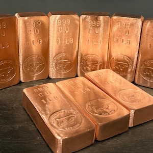 May include: Several copper bars are displayed, each with the markings "999 CU" and "1 LB". The bars have a shiny, reddish-brown color and feature a circular emblem with a bull design.