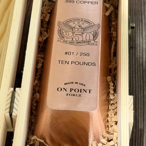 May include: A rectangular .999 copper bar weighing ten pounds, engraved with a 250th anniversary emblem, #01 / 250, and the text Made in USA On Point Forge, nestled inside a light-colored wooden box filled with shredded paper packing material.