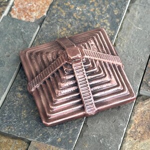 K&ouml;nnte beinhalten: Ein quadratisches, bronzefarbenes Dekorationsobjekt mit pyramidenartigem Design. Das Objekt hat ein riemenartiges Detail auf der Oberseite und besteht aus einem metallischen Material. Das Objekt liegt auf einer Steinoberfl&auml;che.