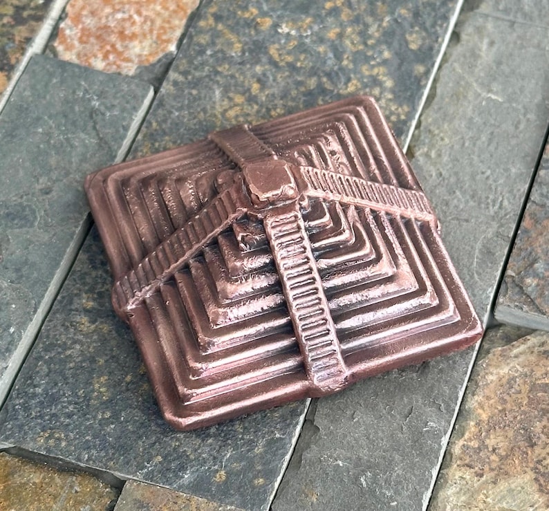K&ouml;nnte beinhalten: Ein quadratisches, bronzefarbenes Dekorationsobjekt mit pyramidenartigem Design. Das Objekt hat ein riemenartiges Detail auf der Oberseite und besteht aus einem metallischen Material. Das Objekt liegt auf einer Steinoberfl&auml;che.