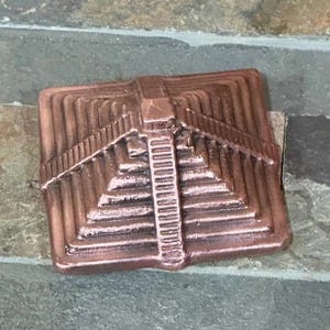 K&ouml;nnte beinhalten: Ein kupferfarbenes, quadratisches Dekorationselement mit einem abgestuften Pyramiden-Design. Die Pyramide hat mehrere Ebenen und eine zentrale Treppe. Das St&uuml;ck ist aus Metall und hat eine strukturierte Oberfl&auml;che.