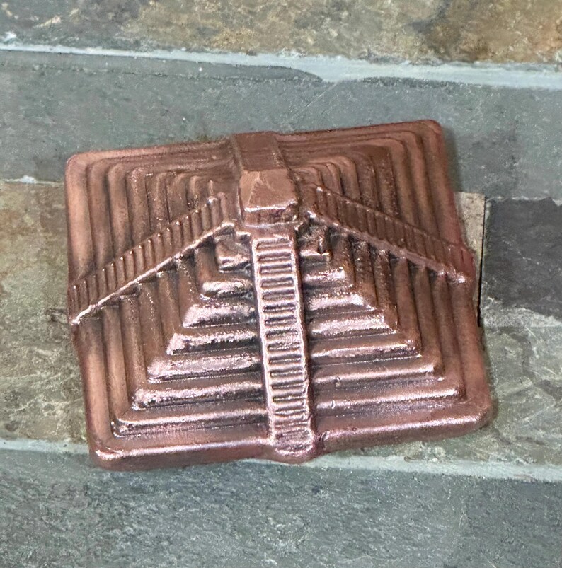 K&ouml;nnte beinhalten: Ein kupferfarbenes, quadratisches Dekorationselement mit einem abgestuften Pyramiden-Design. Die Pyramide hat mehrere Ebenen und eine zentrale Treppe. Das St&uuml;ck ist aus Metall und hat eine strukturierte Oberfl&auml;che.