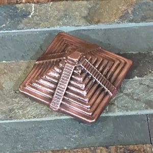 K&ouml;nnte beinhalten: Ein kupferfarbenes, pyramidenf&ouml;rmiges Dekorationsobjekt mit detailliertem, geschichtetem Design. Das Objekt hat ein zentrales, vertikales Element mit einer Reihe horizontaler Linien. Die Pyramide ist quadratisch und sitzt auf einer mehrfarbigen Steinoberfl&auml;che.