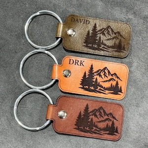 May include: Three leather keychains with silver key rings. Each keychain is rectangular and features a mountain and tree design. The top keychain is olive green and says "DAVID". The middle keychain is orange and says "DRK". The bottom keychain is brown.
