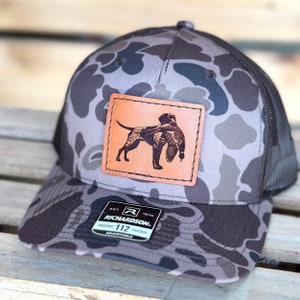 May include: A camouflage trucker hat with a brown leather patch featuring a dog carrying a bird. The hat has a mesh back and a curved brim. The patch is stitched onto the front of the hat. The hat is adjustable.