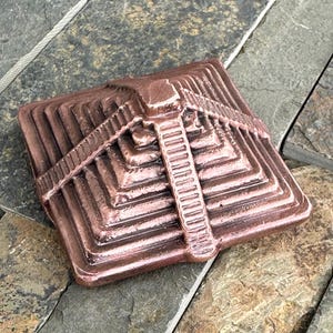 K&ouml;nnte beinhalten: Ein bronzefarbenes, pyramidenf&ouml;rmiges Architekturelement mit einem Stufendesign. Die quadratische Basis erhebt sich in Stufen zu einer zentralen, vertikalen S&auml;ule. Das St&uuml;ck hat ein verwittertes Aussehen, was darauf hindeutet, dass es sich um einen dekorativen Gegenstand f&uuml;r den Au&szlig;enbereich handelt.