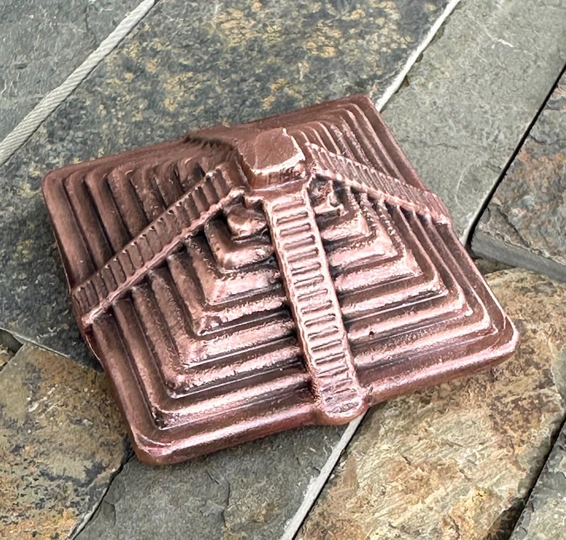 K&ouml;nnte beinhalten: Ein bronzefarbenes, pyramidenf&ouml;rmiges Architekturelement mit einem Stufendesign. Die quadratische Basis erhebt sich in Stufen zu einer zentralen, vertikalen S&auml;ule. Das St&uuml;ck hat ein verwittertes Aussehen, was darauf hindeutet, dass es sich um einen dekorativen Gegenstand f&uuml;r den Au&szlig;enbereich handelt.