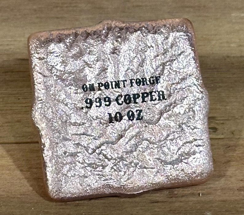 K&ouml;nnte beinhalten: Eine strukturierte, rechteckige Kupferbarren mit dem Text "ON POINT FORGE 999 COPPER 10 OZ" auf der Vorderseite. Der Barren hat eine raue, unebene Oberfl&auml;che und eine metallische, rosafarbene Silberfarbe.
