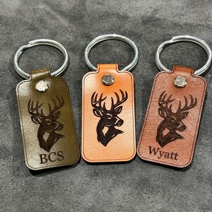 May include: Three leather keychains in shades of brown and orange, each with a silver keyring. The keychains feature a deer head design and personalized text. One keychain reads "BCS", and another reads "Wyatt".