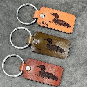 May include: Three leather keychains in shades of brown, each with a silver keyring. The keychains feature a loon design and personalized initials. The top keychain is tan, the middle is olive green, and the bottom is reddish-brown.