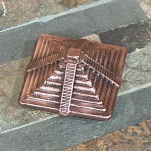 K&ouml;nnte beinhalten: Eine kupferfarbene, quadratische Pyramiden-Skulptur mit detaillierten Stufen und einem zentralen vertikalen Element. Die Metallpyramide hat eine strukturierte Oberfl&auml;che und zeigt ein antikes architektonisches Design. Misst etwa 5 cm in der Breite.