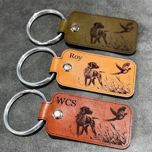 May include: Three leather keychains in olive green, tan, and brown. Each keychain features a dog and bird design, with the text "Roy" and "WCS" on two of them. The keychains have silver metal rings.