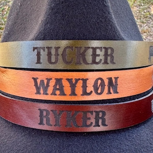 May include: Three leather hat bands in olive green, orange, and burgundy, each with a name engraved in bold letters: "TUCKER", "WAYLON", and "RYKER". The bands are stacked on a black hat, with a small animal silhouette on each end.