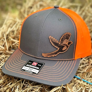 May include: A gray and orange trucker hat with a leather patch of a flying bird. The hat features a gray front panel, an orange mesh back, and orange stitching. The Richardson 112 hat has an adjustable fit.